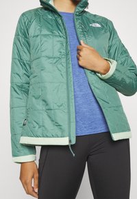 Zelená prošívaná bunda s vysokým límcem a světle zelenými detaily. Má zipový uzávěr a elastické manžety. Nosená s modrým topem a černými kalhotami.