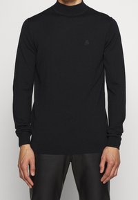 Homme portant un pull à col roulé à manches longues noir uni et un pantalon noir, debout devant un fond clair.