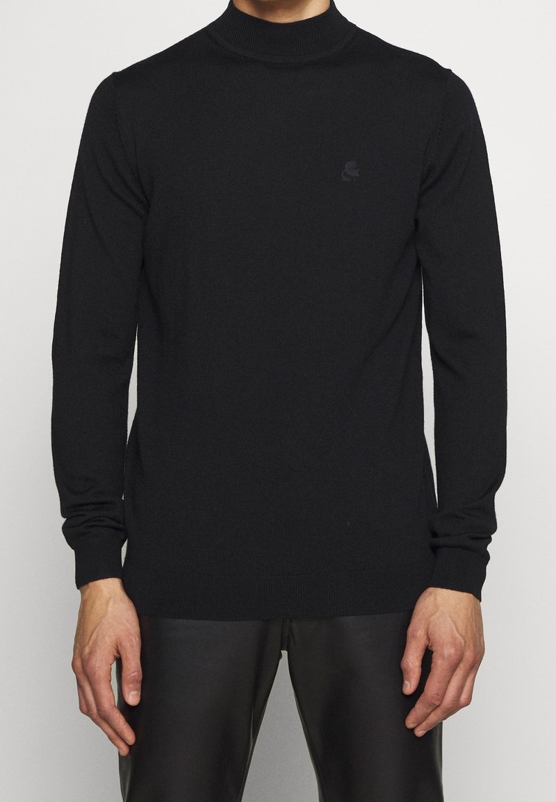 Homme portant un pull à col roulé à manches longues noir uni et un pantalon noir, debout devant un fond clair.