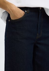 Hand in der vorderen Tasche von dunkelblauen Jeans aus Denim, getragen mit einem weißen Hemd.