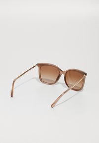 Ochelari de soare supradimensionați, maro, cu un cadru translucid, lentile cu degradé și accente aurii pe tâmplă. Design ușor.