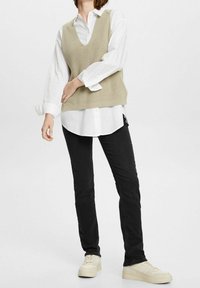 Chaleco de punto beige sobre una camisa blanca de botones, combinado con jeans ajustados negros y zapatillas de color claro. Diseño simple, estilo casual.