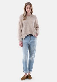 Beige gestrickter Pullover mit V-Ausschnitt, kombiniert mit hellblauen, leicht lockeren Jeans und braunen Slippern mit Kettendetails.