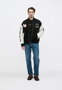 Μαύρο varsity μπουφάν με κρεμ μανίκια, με μπαλώματα και λογότυπα. Φορεμένο πάνω από μαύρο πουκάμισο και συνδυασμένο με μπλε τζιν και καφέ παπούτσια.