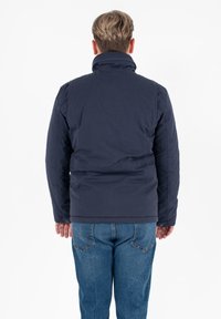 Giacca imbottita blu navy con colletto alto, maniche lunghe e dettaglio con bottoni all'orlo, indossata con jeans azzurri.