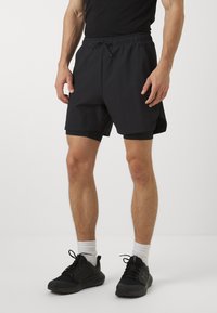 Pantalones cortos deportivos negros con cintura elástica, que incluyen un cordón y un corte ajustado. Llevados sobre pantalones cortos compresivos negros ajustados.