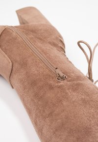 Bottine en daim marron avec fermeture éclair et détail de lacets. Elle présente un talon haut et carré ainsi qu'une texture lisse.
