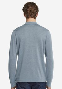 Jasnoniebieska, długorękawowa koszulka polo z kołnierzykiem, gładką teksturą i ściągaczami przy mankietach. Z tyłu widoczny jest subtelny detal na końcu.