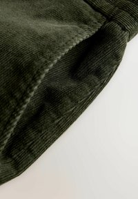 Dark green corduroy fabric displaying textured ridges, folded edge with neat stitching, and soft finish. Close-up view of the material.