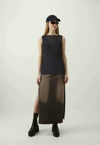 Black sleeveless top paired with a brown, high-slit maxi skirt. Model wears a black cap and dark sunglasses, and chunky black ankle boots.