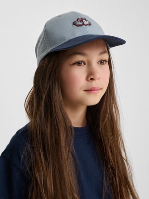 Jong persoon met lang bruin haar, een lichtblauwe pet met een donkere klep en een donkerblauw shirt, kijkt iets naar rechts.