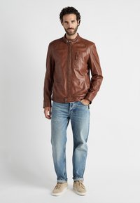 Braune Lederjacke mit Kragen, Vorderreißverschluss und einer Brusttasche. Getragen mit hellblauen Jeans und beigen Sneakern.