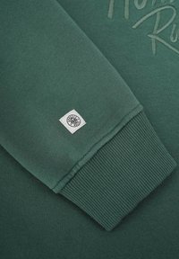 Grön sweatshirt med ribbad mudd, en texturerad logopatch och slät yta, som visar en enkel design och mjukt tyg.