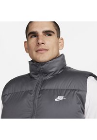 Gilet grigio senza maniche con colletto alto, zip frontale e logo Nike in bianco. Presenta un design trapuntato e una texture liscia.