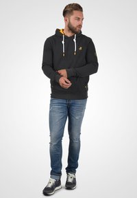 Haut à capuche gris avec doublure jaune, cordons blancs et détails latéraux. Porté avec un jean bleu déchiré et des baskets foncées.