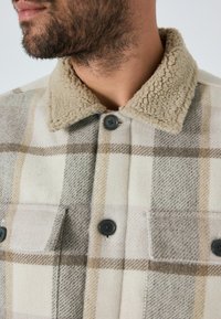 Kariertes Hemd-Jacke mit einem beigefarbenen Schafslederkragen, grauen und braunen Farbtönen, zwei geknöpften Taschen und schwarzen Knöpfen auf einem weichen Stoff.