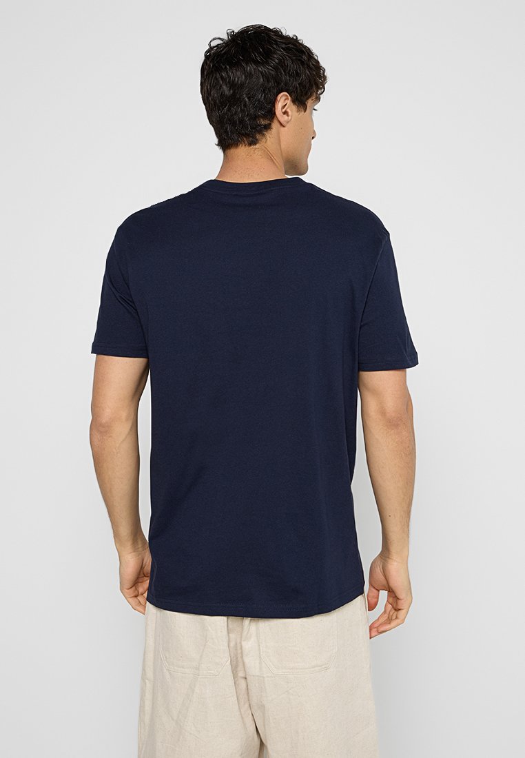 Homme aux cheveux foncés portant un t-shirt simple à manches courtes bleu marine et un pantalon beige, de dos contre un fond clair.