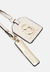 Krémová kožená visačka na zavazadla s černým okrajem, obsahující zlaté logo a kovovou destičku s nápisem "STEVE MADDEN." Texturovaný design, elegantní tvar.
