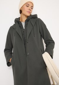 Cappotto verde scuro con colletto a revers, due tasche frontali e un interno grigio in sherpa con zip. Modello che indossa un berretto a maglia crema.
