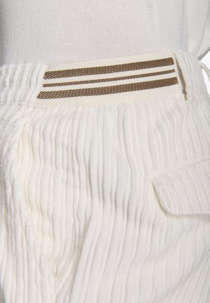 Weiße gerippte Hose mit einer strukturierten Oberfläche, ausgestattet mit einem Bund mit zwei braunen Streifen und einer seitlichen Tasche für zusätzliche Details.