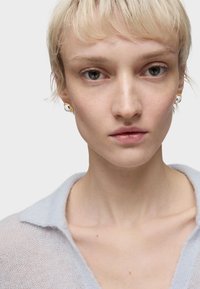 Modello che indossa un maglione leggero azzurro chiaro in tessuto a rete con colletto. Orecchini circolari dorati e neri visibili. Pelle liscia, capelli biondi corti.