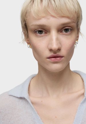 Modello che indossa un maglione leggero azzurro chiaro in tessuto a rete con colletto. Orecchini circolari dorati e neri visibili. Pelle liscia, capelli biondi corti.
