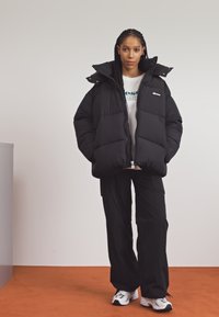 Čierna puffa bunda s vysokým golierom, kombinovaná s bielym tričkom, čiernymi cargo nohavicami a bielymi teniskami. Hladká textura, oversized strih.