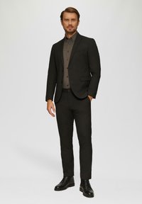 Schwarze Sakko und taillierte Hose über einem karierten Knopfhemd. Glatter Stoff, maßgeschneiderte Passform, mit schwarzen Lederstiefeln. Einheitliche Farbe mit dezenten Mustern.