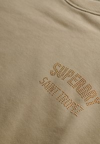 Sudadera beige de tejido suave, con "SUPERDRY" y "SAINT TROPEZ" en tachuelas metálicas, cuello redondo y superficie texturizada.