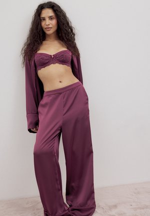 Moteris dėvi slyvinės spalvos satino platėjančias kelnes, derančią nėriniuotą liemenėlę ir atvirą švarką, stovinti priešais paprastą šviesiai pilką foną.