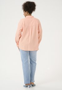 Camisa de manga larga rosa claro con pliegue en la espalda y dobladillo redondeado, combinada con pantalones vaqueros rectos azul claro y zapatos de tacón azules.