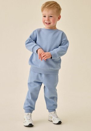 Smilende småbarn iført en lyseblå sweatshirt og matchende bukser med hvide sneakers, stående mod en ensfarvet beige baggrund.