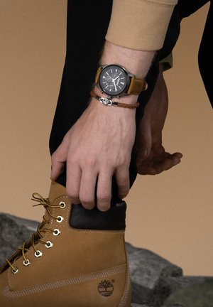 Botte en cuir marron avec col noir et œillets dorés, associée à une montre-bracelet avec un cadran sombre et un bracelet en cuir marron.