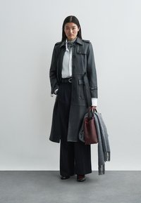 Long black leather coat with button closure, paired with a white shirt and navy high-waisted trousers. Accessories include a burgundy bag and grey scarf.