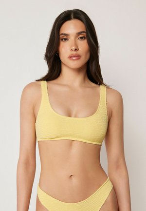 Kvinde med langt mørkt hår iført en tekstureret gul bikini overdel og underdel, stående mod en ensfarvet lys baggrund.