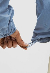 Vaaleansininen takki, jossa on joustavat hihansuut ja nyörit hihoissa. Materiaali näyttää kevyeltä ja sileältä.