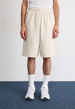 Beige bavlněné kraťasy s elastickým pasem, bočními kapsami a volným střihem, v kombinaci s bílými sportovními ponožkami a teniskami.