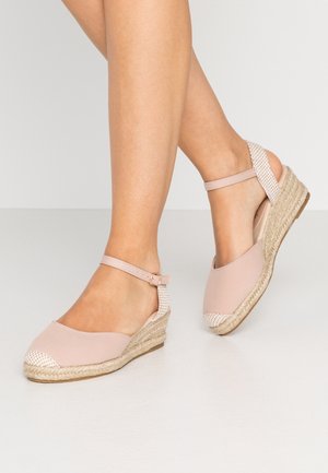 Ben iført lyserøde kile-espadrilles med ankelremme og vævede detaljer på hæl og tå, stående på en glat hvid gulv.