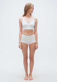 Νέα γυναίκα όρθια, ξυπόλυτη, φοράει λευκό κοντό τοπ και σλιπ ψηλόμεσο, κοιτάζει προς το πλάι μπροστά σε απλό φόντο.