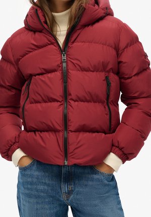 Personne portant une doudoune rouge avec des fermetures éclair noires sur un pull blanc et un jean bleu, les mains dans les poches de la veste.