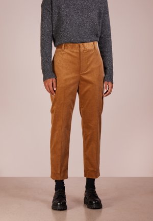 Persoon die bruine ribfluwelen broek, zwarte sokken, zwarte schoenen en een donkergrijs gemêleerde wollen trui draagt en staat op een lichte vloer.