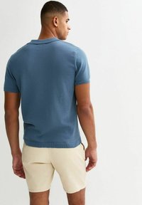 Camisa polo de punto azul de manga corta, con tejido texturizado, cuello clásico y costuras reforzadas, usada con pantalones cortos beige, vista trasera.