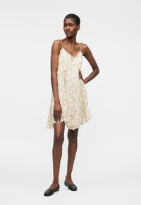 Femme debout, main sur la hanche, portant une robe blanche sans manches à fleurs et des chaussures plates noires sur un fond clair uni.