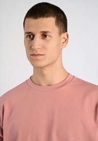 Ljusrosa bomulls t-shirt med rund hals, visad på en modell. Tyget ser slätt ut med en enkel, odekorerad design.