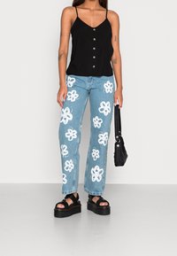 Camisole noire avec boutons sur le devant, jeans bleu clair avec des motifs de fleurs blanches, et sandales à plateforme noires avec un sac à main noir.