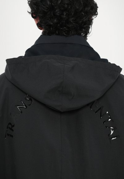 Jachetă neagră cu glugă, cu cuvântul "TRANS" decupat într-o finisare lucioasă pe spate. Textură netedă; design minimalist.