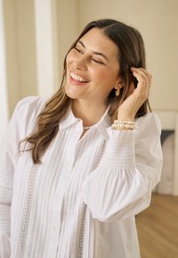 Weißes Hemd zum Zumachen mit Falten und Spitze. Lange, aufgeblähte Ärmel mit gerafften Bündchen. Getragen mit Perlenarmbändern und goldenen Ohrringen.