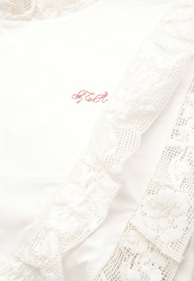 Άσπρο ύφασμα με λεπτή φλοράλ δαντέλα και διακριτικά κεντημένα κόκκινα γράμματα που διαφαίνονται εν μέρει κάτω από τη δαντέλα.