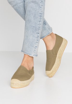 Fødder iført olivengrønne slip-on espadrillos med flettede jutesåler og lyseblå cropped jeans med flossede kanter mod en hvid baggrund.