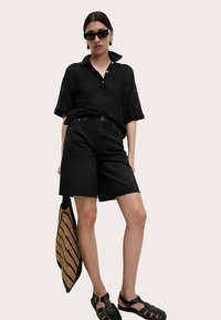 Svart stickad pikétröja med korta ärmar, högmidjade svarta shorts, svarta sandaler och en vävd strandväska med randigt mönster.